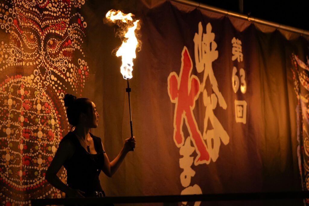 A fire performer holding a torch against a backdrop of Japanese festival art at night.